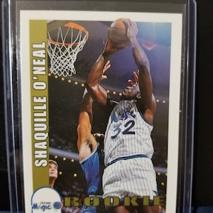 May include: A basketball trading card featuring Shaquille O'Neal in a blue and white Orlando Magic uniform, with the text "Shaquille O'Neal" and "Rookie" on the card.