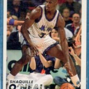 May include: A basketball card featuring Shaquille O'Neal, a professional basketball player, wearing a white and purple Orlando Magic jersey. The card is from the 1993-94 Fleer series.