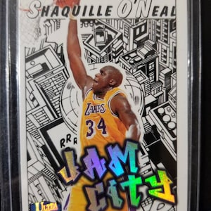 May include: A basketball trading card featuring Shaquille O'Neal in a purple and gold Los Angeles Lakers uniform, dunking a basketball. The card has a black and white background with a city skyline design and the words "Slam City" in a holographic font.