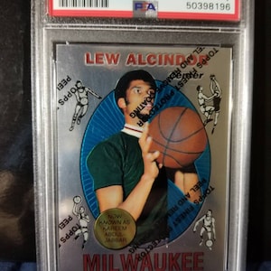 May include: A 1996 Topps Finest Reprint basketball card featuring Lew Alcindor, now known as Kareem Abdul-Jabbar, in a green jersey with a basketball in his hands. The card is in a protective case with a PSA grading label.