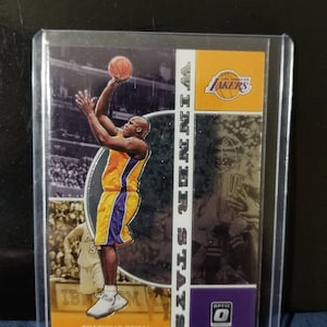 May include: A basketball trading card featuring Shaquille O'Neal in a purple and gold Los Angeles Lakers uniform. The card is from the Optic series and features a silver foil background with a collage of images.