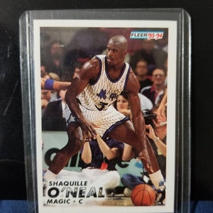 May include: A trading card featuring a basketball player in a blue and white uniform. The player is Shaq O'Neal, a centre for the Orlando Magic. The card is from the 1993-94 Fleer series.