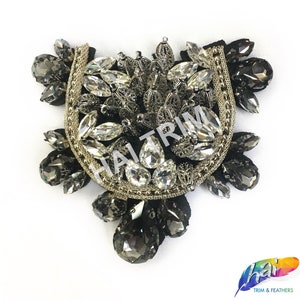 Puede incluir: Una aplicación de cuentas plateadas y negras con forma de media luna y adornos de cristal. La aplicación está sobre un fondo de tela negra.