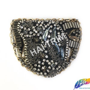 Puede incluir: Una aplicación de tela negra con una variedad de adornos, incluyendo pedrería, cuentas y cadenas metálicas. La aplicación tiene la forma de un óvalo curvo.