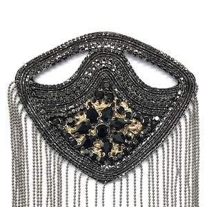 Puede incluir: Una decoración de cuentas negras y doradas con una franja de cadenas plateadas. La decoración tiene forma de lágrima y tiene un diseño floral en el centro.