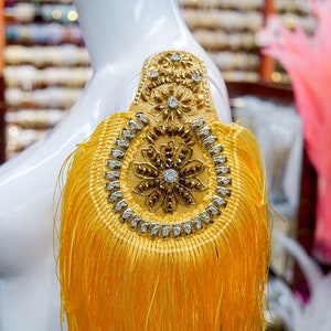 Puede incluir: Un ribete de flecos amarillo con un adorno de oro y pedrería. El ribete está en un maniquí blanco y probablemente se utiliza para el diseño de vestuario o moda.