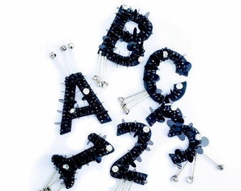 May include: Black sequined letters A, B, C, 1, 2, and 3 with silver pins. The letters and numbers are sewn with black sequins and have a shiny finish. The pins are attached to the back of each letter and number.