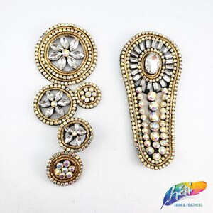 Op de afbeelding: Twee goudkleurige strass-versieringen voor kleding of knutselen. De ene is een cluster van cirkels met een bloemmotief in het midden. De andere is een lange, smalle vorm met een enkele grote strass-steen in het midden.