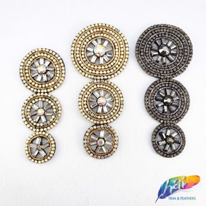 May include: Three sets of decorative embellishments, each with three circular pieces. The top piece is the largest, followed by a medium-sized piece, and then the smallest piece. The embellishments are in gold and silver tones with rhinestones.