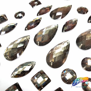 May include: Assortment of iridescent, faceted, glass rhinestones in various shapes, including teardrop, oval, and square. The rhinestones have a dark brown, almost black, color.