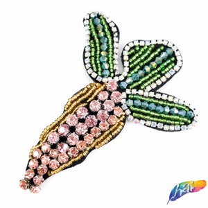 May include: A green and pink beaded carrot with a black backing. The carrot is made of small beads and rhinestones.