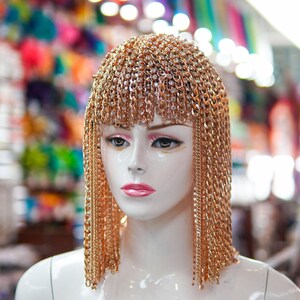 Op de afbeelding: Een witte mannequinkop met een gouden kettinghoofdtooi met strass. De hoofdtooi is gemaakt van meerdere strengen ketting die aan de zijkanten van het hoofd hangen. De mannequin staat voor een wazige achtergrond van kleurrijk textiel.