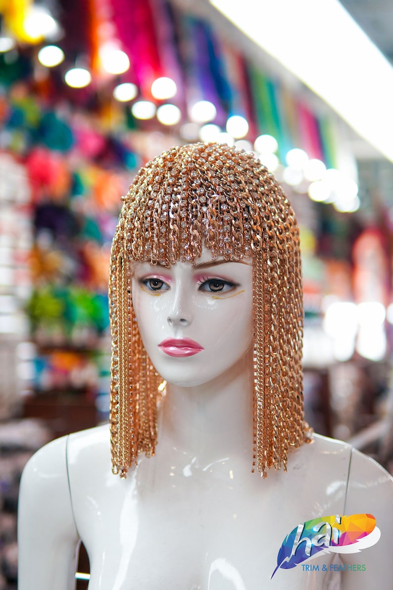 Op de afbeelding: Een witte mannequinkop met een gouden kettinghoofdtooi met strass. De hoofdtooi is gemaakt van meerdere strengen ketting die aan de zijkanten van het hoofd hangen. De mannequin staat voor een wazige achtergrond van kleurrijk textiel.