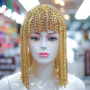 Op de afbeelding: Een gouden kettinghoofdtooi met strass, gedragen door een wit mannequin. De hoofdtooi is gemaakt van meerdere strengen gouden ketting met strass eraan bevestigd. De hoofdtooi is gestyled om eruit te zien als een pruik.