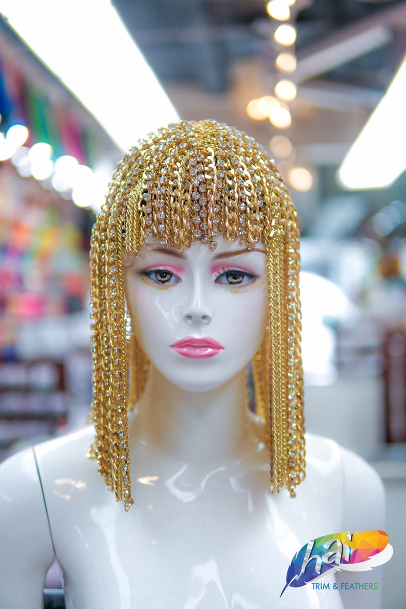 Op de afbeelding: Een gouden kettinghoofdtooi met strass, gedragen door een wit mannequin. De hoofdtooi is gemaakt van meerdere strengen gouden ketting met strass eraan bevestigd. De hoofdtooi is gestyled om eruit te zien als een pruik.