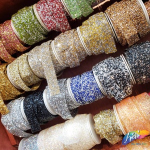 May include: A variety of sparkly rhinestone trim rolls in different colors, including gold, silver, black, blue, and pink. The rolls are arranged in a display on a wooden surface.