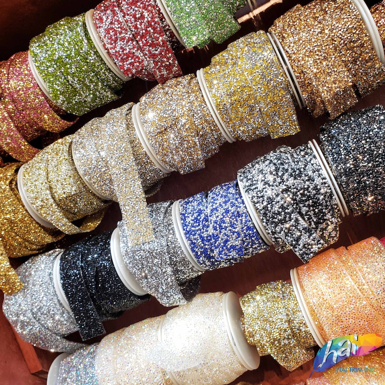May include: A variety of sparkly rhinestone trim rolls in different colors, including gold, silver, black, blue, and pink. The rolls are arranged in a display on a wooden surface.