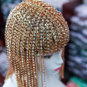 Op de afbeelding: Een gouden ketting haarstuk met strass. Het haarstuk is gemaakt van veel kleine kettingen die aan elkaar zijn verbonden. De kettingen zijn versierd met kleine strass steentjes. Het haarstuk wordt gedragen op een mannequin hoofd.