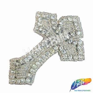 May include: A silver cross-shaped embellishment with clear rhinestones. The cross is made of metal and has a shiny finish.