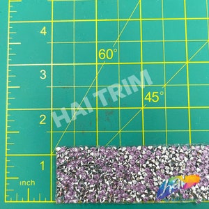 May include: A close-up of a silver and purple rhinestone trim, measuring approximately 1 inch in length. The trim is on a green cutting mat with markings for inches and angles.
