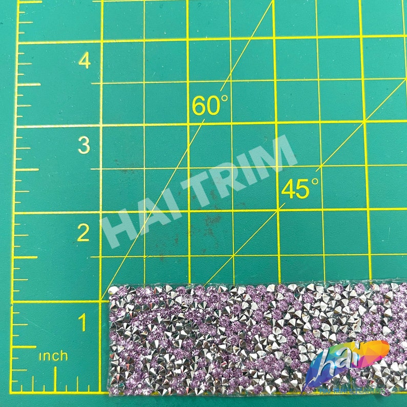 May include: A close-up of a silver and purple rhinestone trim, measuring approximately 1 inch in length. The trim is on a green cutting mat with markings for inches and angles.