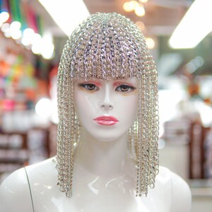 Op de afbeelding: Een witte mannequin-kop met een zilveren kettingmail-hoofdtooi met lange strengen ketting die aan de zijkanten naar beneden hangen. De mannequin staat in een winkel met een bord waarop "hai trim & feathers" staat.