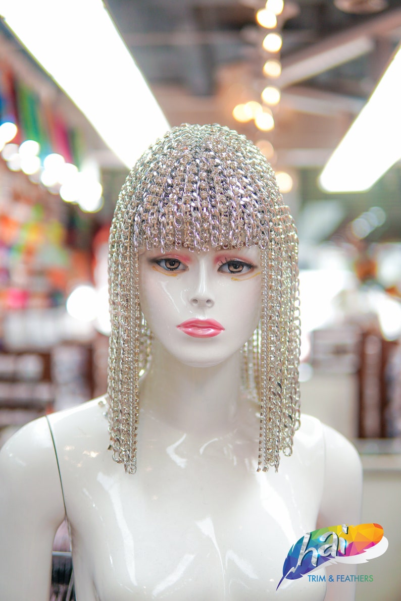 Op de afbeelding: Een witte mannequin-kop met een zilveren kettingmail-hoofdtooi met lange strengen ketting die aan de zijkanten naar beneden hangen. De mannequin staat in een winkel met een bord waarop "hai trim & feathers" staat.