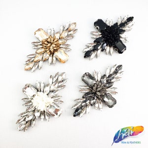 May include: Four different cross-shaped embellishments with rhinestones and a flower in the center. The flowers are in black, white, gray, and gold.