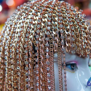 Op de afbeelding: Een gouden kettinghoofdtooi met strass. De hoofdtooi is gemaakt van meerdere kettingen die aan elkaar zijn verbonden. De kettingen zijn versierd met kleine, heldere strasssteentjes.