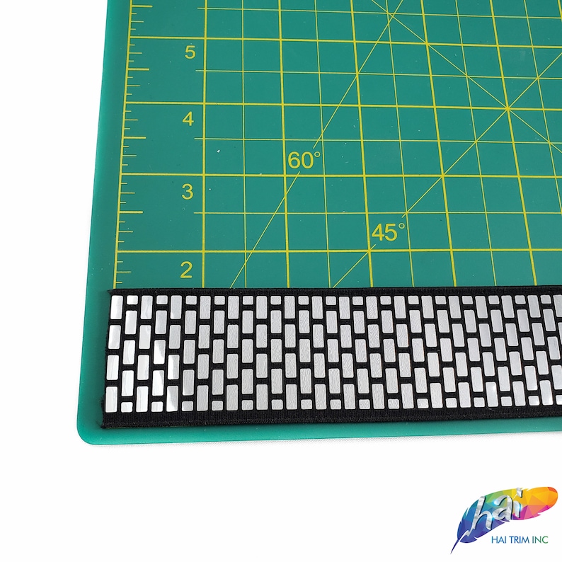 May include: Black and silver elastic trim with a geometric pattern. The trim is shown on a green cutting mat with markings for 45 and 60 degrees.