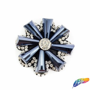 Puede incluir: Una decoración en forma de flor de color azul oscuro con un gran strass transparente en el centro. La flor está rodeada de strass más pequeños.