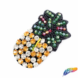 May include: A black fabric pineapple patch with green, red, and yellow rhinestones. The pineapple is made of beads and rhinestones.