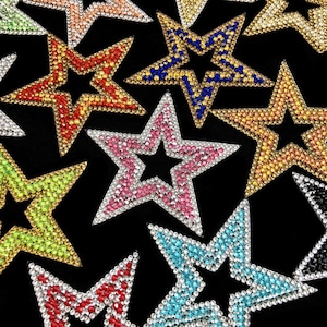 May include: A collection of star-shaped patches covered in small, sparkling rhinestones in various colors including pink, blue, yellow, red, green, and orange, arranged against a solid black background, each featuring a hollow center.