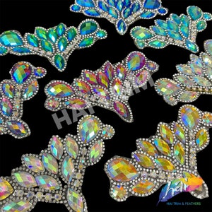 May include: Close-up of several decorative rhinestone appliques in various colors, including blue, green, and iridescent. Each applique features a leaf-like design with multiple crystal-like stones and a silver-colored backing. The appliques are arranged on a black background.