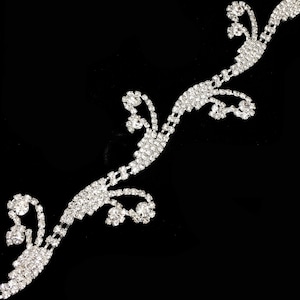 May include: A sparkling silver rhinestone trim with a floral vine design. The trim is set against a black background, showcasing the intricate details of the rhinestones. This decorative trim is suitable for various craft projects.