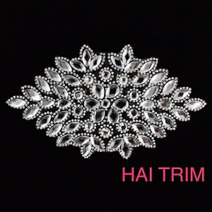 May include: A clear rhinestone applique with a floral design. The applique features various shapes of rhinestones, including oval and round, arranged in a symmetrical pattern. The text "HAI TRIM" is displayed in pink.