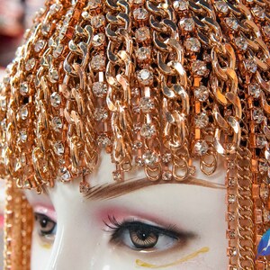 Op de afbeelding: Een goudkleurig hoofdtooi met een kettingontwerp en heldere strass steentjes. De hoofdtooi wordt gedragen door een mannequin met een wit gezicht en bruine ogen.