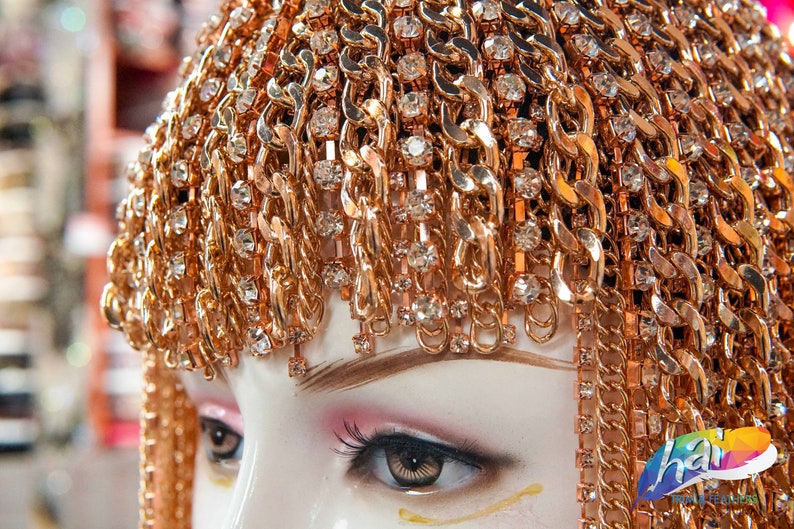 Op de afbeelding: Een goudkleurig hoofdtooi met een kettingontwerp en heldere strass steentjes. De hoofdtooi wordt gedragen door een mannequin met een wit gezicht en bruine ogen.
