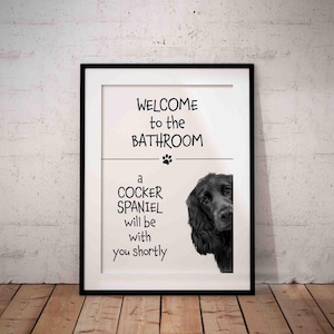 May include: Framed print with a black border. The text reads "WELCOME to the BATHROOM" above a paw print and a line. Below, it says "a COCKER SPANIEL will be with you shortly." A black Cocker Spaniel peeks from the right.