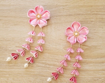 May include: Two pink fabric flower hair accessories with dangling petal accents and pearl beads. The flowers have a central pearl and gold accents. The accessories are set against a light wood grain background.