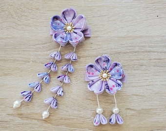 May include: Two lavender fabric flower hair clips. One clip features a cascading design with pearl accents, while the other has two dangling pearl accents. The flowers have a star pattern in the center and are set on a light purple clip.