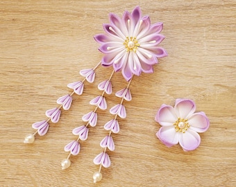 May include: Two handmade, lavender and white fabric flower hair accessories. One large flower with cascading lavender petals and pearl accents, and a smaller matching flower. The flowers are on a light wood surface.