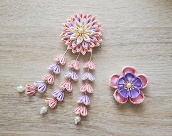 May include: Two handmade fabric flower hair accessories. One is a large, multi-layered flower with cascading petal strands, in shades of pink, lavender, and cream, adorned with pearls. The other is a smaller, five-petal flower in pink and purple.