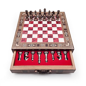 Op de afbeelding: Een houten schaakset met een rood-wit geblokt bord en een lade met schaakstukken. Het bord is omlijst met decoratief inlegwerk. De schaakstukken zijn zilver en brons.