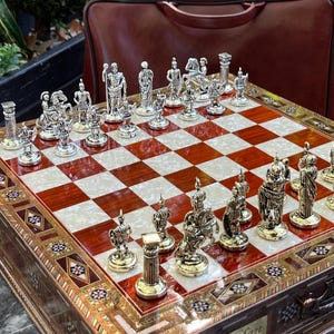 May include: A wooden chessboard with a red and white chequered playing surface. The chess pieces are silver and feature a variety of designs, including knights on horseback, rooks, and bishops. The chessboard is inlaid with intricate patterns of wood and mother of pearl. The chessboard is in a wooden box with a brass latch and the inscription "Turqut Ceylan".