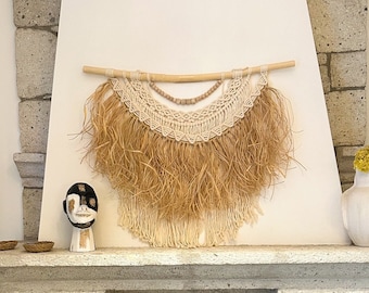 Könnte beinhalten: Ein beige-weißer Makramee-Wandbehang mit einem Holzstab und Fransen. Der Behang ist über einem Steinkamin mit einem Holzmantel angebracht.