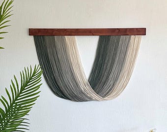 May include: A wall hanging with a wooden top and cascading strands in shades of gray and cream. Below, a wooden bench holds cream-colored pillows with tassels and ruffles. A potted plant and a vase of flowers complete the scene, creating a cozy, neutral-toned aesthetic.