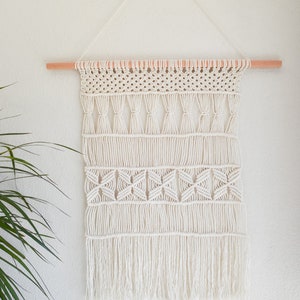 Könnte beinhalten: Ein weißer Makramee-Wandbehang mit einer Holzleiste oben. Der Behang hat ein geometrisches Muster mit Fransen unten.