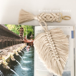 May include: A beige macrame leaf keychain with a tassel and a gold metal key ring. The keychain is hanging from a book with the text "BALI AND LOMPOK AREA BY AREA 83" and "CENTK" visible.