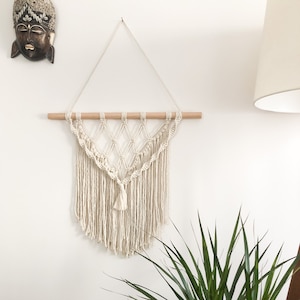 Peut inclure: Une suspension murale en macramé blanche avec une baguette en bois en haut. La suspension a la forme d'un triangle avec une frange en bas. La suspension est sur un mur blanc avec une plante verte au premier plan.